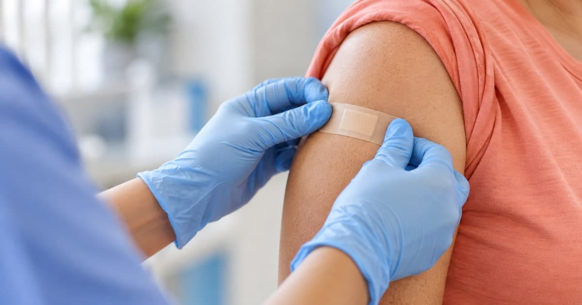 Gloved nurse applying a small bandage to a patient’s upper arm after vaccination.